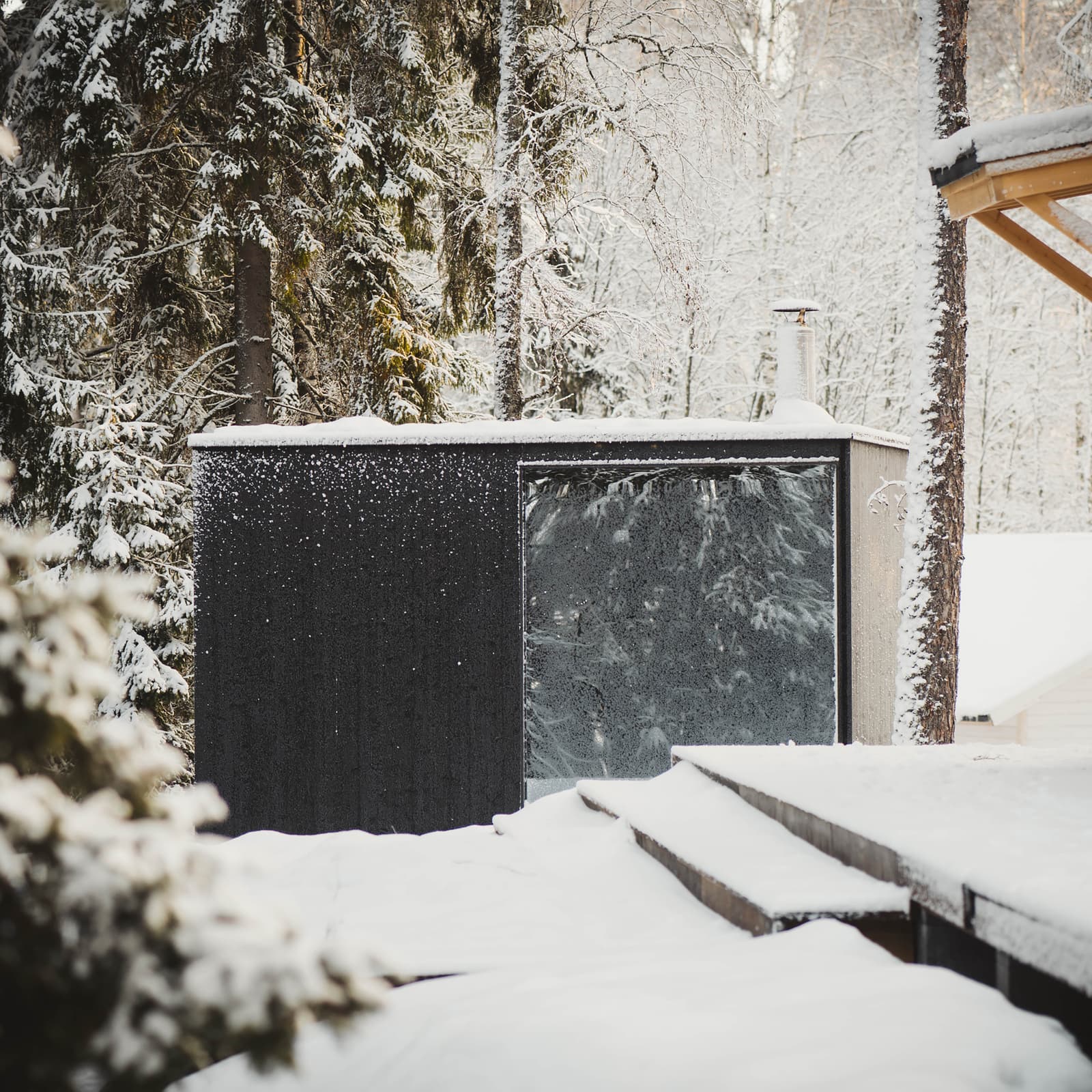 A modern, outdoor sauna with a black facade and a large, mirrored window stands in a snowy forest next to a wooden terrace.