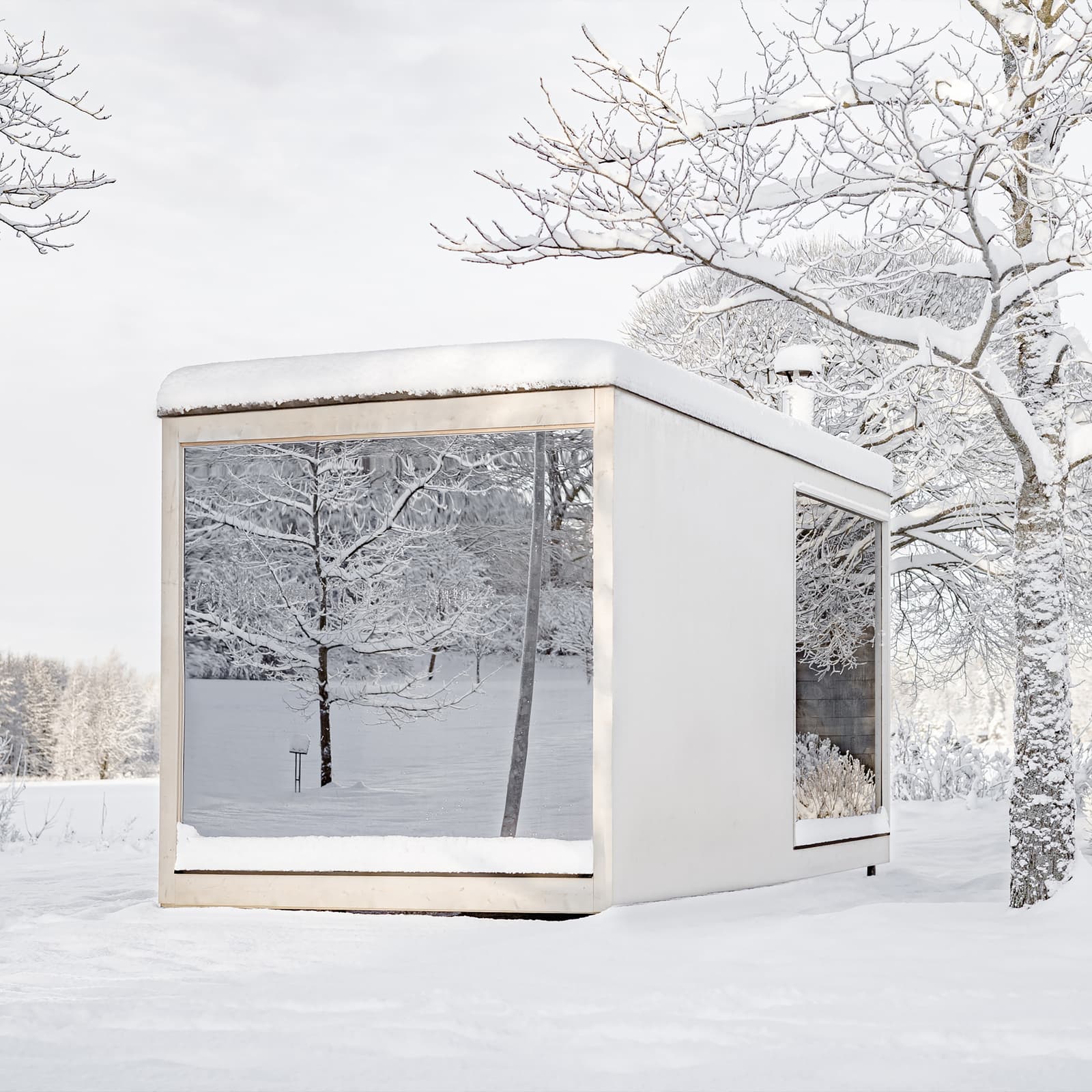 A white, minimalist cabin with panoramic windows stands in a snow-covered garden. The snow-covered trees are reflected in the panes.